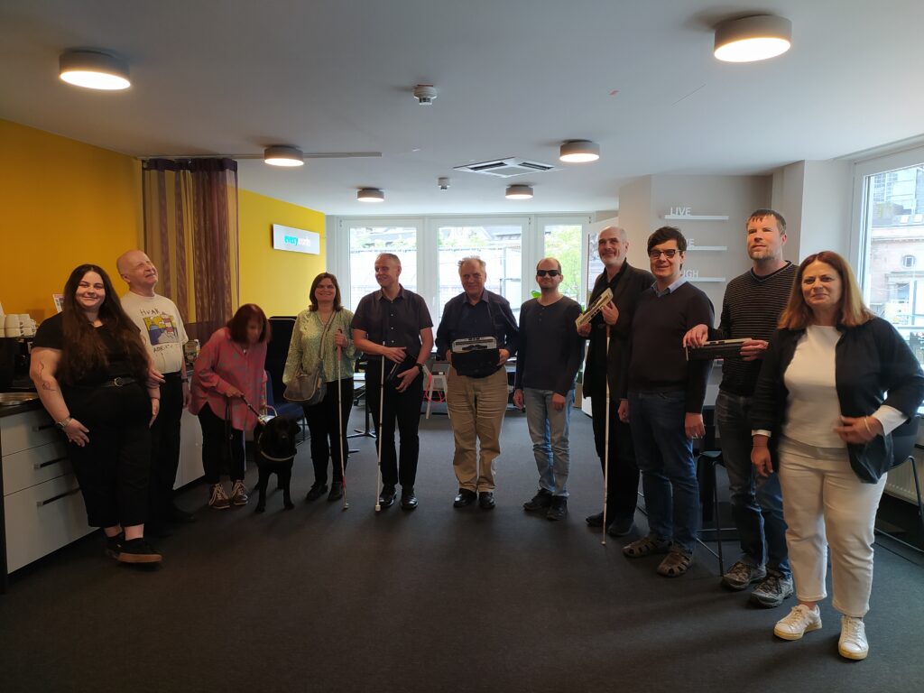 A photo of the Working Group members: This photo captures a group of people standing together in a room. The individuals are standing side by side, smiling, and seem relaxed. Some members of the group are holding tactile writing devices, possibly Braille devices or tools, while a couple of people are using white canes. One person is accompanied by a guide dog. The room has yellow walls and large windows in the background that allow natural light to fill the space. The decor is modern. There are shelves with possibly motivational phrases. The mood of the image is friendly and inclusive, with the group appearing to enjoy each other’s company. The setting seems like an accessible workspace or meeting venue, likely accommodating the needs of visually impaired participants.