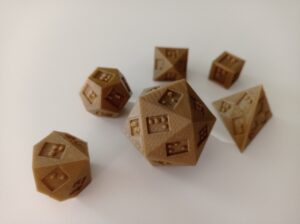 Six different polyhedral dice with braille numbers laying on a white surface. Each dice has a different number of sides: In the front there is one die with 10 faces, one with 20 faces, and a third one with 4 faces with the shapes of all faces being triangles. Behind these thera are 3 dice with 12, 8, and the standard 6 faces respectively. The 8 sided die has triangular faces too while the 12 faced die is compiled of pentagons. The dice all have a caramell-brown colour and the lined texture of 3-D prints.
