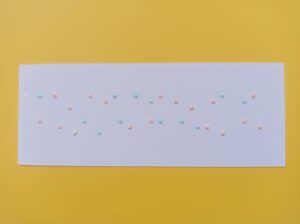 A white piece of paper on a yellow background. On the paper there are “deconstructed” braille letters with pink and light blue dots meant to form the name “Andrea Schmidt” in two lines. Each dot is written as if it were its own letter so that the dots form two abstract lines of dots.