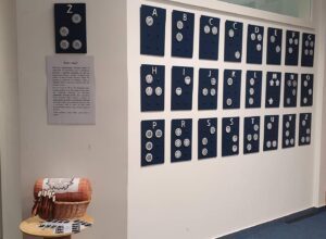 The Slovenian braille alphabet made out of circles of lace exhibited in a museum. Each dot is represented by a circle of approximately 20 centimetres in diameter made from white threat. The dots for each letter share a different floral pattern. Each letter is stitched onto a black rectangular board together with the corresponding roman letter. The letters hang on a wall with a description of the piece in Slovene next to them. Infront of the description, there is a small round table with exemplary lacemaking tools propped up on top: A tube shaped basket with the blueprint of a lace design attached as well as a number of wooden spules with white threat.