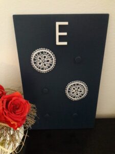 The braille letter e made from white lace on a black background. The braille dots are designed in a shape resembling a blossom with 5 petals. Above the braille letter there is a white roman letter E.  On the left of the letter there is a red rose.