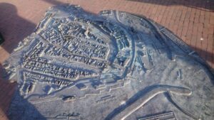 A picture of the entire tactile model of the Harlingen city centre. The model is made out of bronze in a scale of 1:1000 with a size of roughly 80 by 120 centimetres. The outlines are matching the shape of Harlingen. All buildings as well as harbour structures and vessels are modelled. The names of landmarks and canals are written in both tactile roman letters and braille.