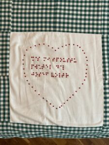 A picture of a blanket made of green and white checkered fabric laying on a table. In the middle of the blanket is a white square embroidered with red beads forming the shape of a heart. The heart is filled with a line from the Czech song Krásný Čas by Josef Voltr written in braille made from beads: “Čas mi spóusťu přáťel dal – Jósef Vólťr” – “Time has given me many friends”.