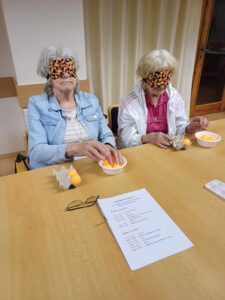 The participants with the highest age in the camp with their eyes covered (aged 78). They are sitting at a table and having fun learning how to find their way around a six-point compass using balls that they place in a six-pointed egg carton.
