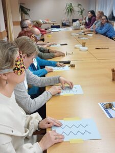 Participants sitting at a table. Some have their eyes covered. Each has a relief sheet in front of them with various shapes and lines, which they have to trace with their fingers and describe in words.