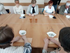 Participants sitting at a table. Some have their eyes covered. Each has a pinboard in front of them. Following instructions, they write letters of the Braille alphabet on it.
