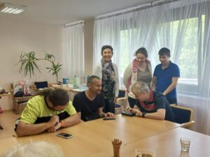 In the photo at the table, people from Touch&Speech n. o. explain and demonstrate how to play Braille Tetris on a Braille display. One participant tries the game at the table and comments on it. Three others stand around, watching and laughing at the game and the comments.