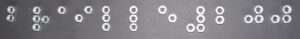 Auf einem Untergrund liegen Schraubmuttern,
die gemeinsam in Brailleschrift den Schriftzug „Braille 200“ bilden.