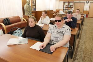 The photograph depicts people sitting in a room resembling a library. In the foreground, there are two women (Svetlana and Julia); Svetlana is wearing sunglasses and is touching a tactile surface, as if reading Braille. Svetlana uses a plastic slate with 27 lines, 30 cells each. Julia uses a metal slate with 18 lines, 24 cells each. Other participants behind them, including a woman and two men, sit attentively. On the tables in front of them, there are books and materials. In the left corner, there's a sculpture, and along the wall are shelves with books. The atmosphere in the room is calm and focused.