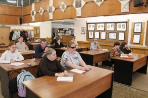 The image depicts a room setting where several individuals are seated at wooden desks. The room has a traditional classroom layout with two rows of desks. Most people are writing, and they appear to be focused and attentive. The individuals are diverse in appearance, and their attire is casual. The walls of the room are adorned with various framed artworks and documents, adding an academic feel to the space. Above the framed items, there are decorative geometric patterns along the wall. In the background, there are open bookshelves filled with books, and several coats are hanging on a coat rack. The lighting in the room is bright, contributing to a focused atmosphere. The overall ambience is that of a study session or a lecture in progress.