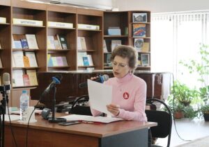 The image shows the director of the library Elena Zakharova sitting at a table. She is reading from a sheet of paper and holding a microphone with a blue cover. She is wearing a pink turtleneck and glasses. On the table in front of her are two microphones on stands, a bottle of water, and several items, including a phone and a pencil. Behind her, there are shelves filled with books and documents. The room also features a piano and several potted plants.