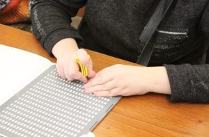 The photo shows a person's hands using a stylus for writing on a Braille slate. The person is holding a yellow stylus and is pressing on a sheet of paper with Braille characters, which consist of small, raised dots arranged in patterns. The metal slate is on a wooden table. The person is wearing a dark long-sleeved top with subtle glittery details and a zippered pocket. A strap is visible over the shoulder, suggesting they might be carrying a bag.
