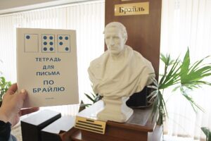 The image shows a person holding a notebook. The cover features Braille alphabet depicted as blue dots, and the text reads in Russian: "Notebook for writing in Braille" In the background, there is a white bust of Louis Braille draped in classical attire. Above the bust, a plaque reads in Russian "Braille." There are green plants on either side of the bust, which is mounted on a wooden stand. The room is well-lit, likely due to light coming through vertical blinds on the windows.