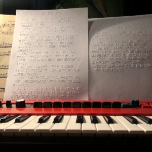 The image shows a close-up of the keys of an organ. Two pages of Braille music notation lie on the music stand, and on the left is an excerpt from an old standard score. The score shows a segment of Johann Sebastian Bach's Two-Part Invention in E major.