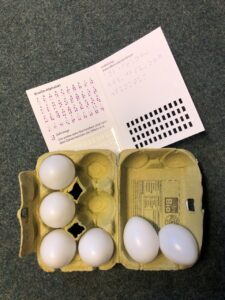 The photo shows a six-egg cardboard box lying on a blue-grey carpet. Four white plastic eggs are arranged to represent the Braille letter „F“, while the remaining two eggs are placed on the lid of the box. Above the egg box, there is a small flyer from the DBSV featuring the Braille alphabet.