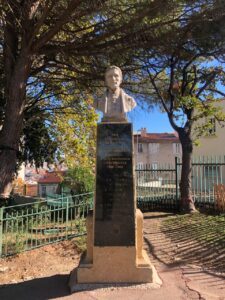 The first photo features a statue in honor of Louis Braille. The statue is located in the garden of Colline Puget Park, in the city center of Marseille, in the south of France. Dating from 1952, the statue is a monument dedicated by the blind population of Provence to Louis Braille. The white stone bust rests on a stone column with a dark grey marble base that bears an inscription in gold lettering and Braille script, as well as a drawing of a hand. Behind the statue stands a large pine tree, and further back are fences enclosing the park, with some houses in the background.