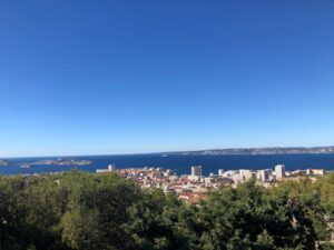 The photo features the view from Puget Park, located on a hill in the city center of Marseille. Near the statue of Louis Braille, trees occupy the foreground, the city extends into the distance, the nearby harbor borders the Mediterranean Sea, and the small Frioul Islands lie just off the coast under a clear blue sky.