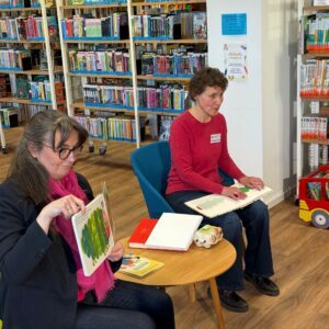 The photo features two people in the Quickborn public library. Giuseppina Dolle is seated on a blue chair with a children’s book on her lap that includes illustrations, printed text, and Braille. She reads the Braille with her fingers. To her right sits Kerstin Kranz, who is holding a book and showing its illustrations to the audience. Between them is a small round oak table holding several books and an egg box. Large bookshelves fill the background.