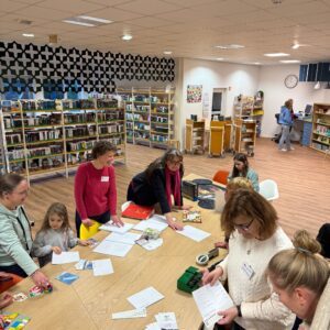 The photo features the Quickborn public library. It highlights a large rectangular wooden table on which there are several Braille-related objects, such as books, flyers with the Braille alphabet, a six-egg carton, Uno, card games, a puzzle, and a small Braille typewriter. Around the table are several people, adults and children, some sitting and others standing. The mood appears joyful, as some people are smiling. In the background, there are large bookshelves.