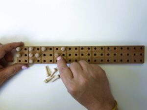 A person forms the letters B, R, A, I with wooden pins on the wooden Braille board.