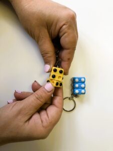 Two small Braille keychains: a person holds a yellow one with black dots and turns it, while a blue one with white dots lies on the table.