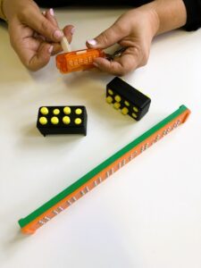 Three types of slates with pressable Braille dots: one orange and green with silver dots and space for 12 letters, two identical black plastic slates with 8 yellow dots each, and one small orange keychain slate. A person writes on the small slate with a stylus.