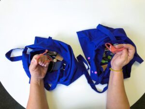 	On a white table, there are two identical blue tote bags containing different objects. Hands take one item from each bag and reveal that the identical items found are two jar rubber seals.