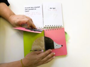 The book is open and shows the following:
Top left: Autsch! Was stachelt und raschelt im Laub? in print and Braille
Top right: der Igel in print and Braille
Bottom left: a hand flips the page to show the back of the animal
Bottom right: the front of the hedgehog