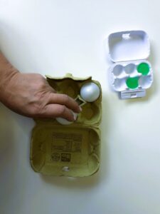 A 6-egg paper carton has two white plastic eggs. A person places eggs in positions 1 and 5 to form the letter E. Next to it, a smaller white quail egg carton has two green rubber balls also arranged like the letter E.
