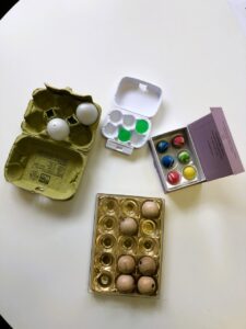 On a table, the two egg cartons are accompanied by praline boxes. A small one with 6 slots is filled with colorful rubber balls. A Ferrero Rocher plastic box with 15 slots shows the letters E and G in Braille.