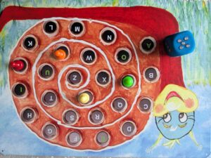 In this image, the snail illustration once again displays the alphabet in printed text. A die with raised numbers is shown nearby, and colored pins are placed on some of the letter pieces: a red pin on J, a green pin on P, an orange pin on V, and a yellow pin on Y.