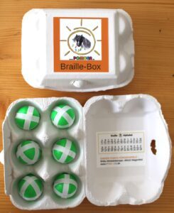 The photo features two six-egg cartons on a table. The carton on top is closed and has a sticker with Pomuga’s logo. The other box is open and contains six green-and-white balls instead of eggs. On the lid, there is the Braille alphabet.