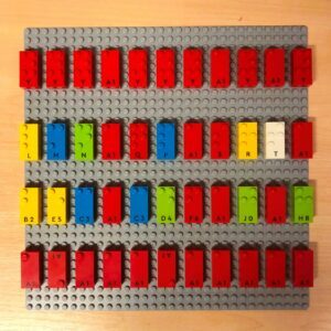 The gray LEGO plate has LEGO bricks with Braille letters on them.
1st row:
Y Y Y A Y Y Y A Y A Y
2nd row:
L M N A Q P A S R T A
3rd row:
B E C A C D F A J A H
4th row:
A row of 11 A's. The 2nd and 6th A's are upside down.