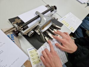The photo shows Hands on the keys of a braille machine, a braille alphabet lying under the hands shows the dot combinations.