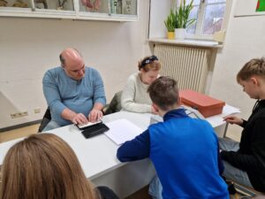 The photo shows Nermin and Anja are sitting at a table in the classroom, opposite three students at braille machines.